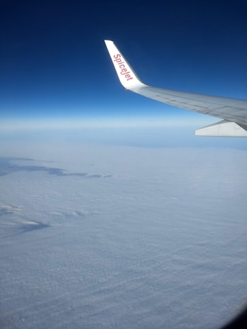 A clear view from an airplane window showing the wingtip of an aircraft with the SpiceJet logo against a backdrop of a vast cloudscape and deep blue sky.