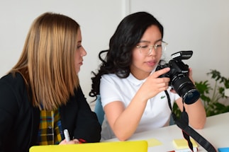 Two people are sitting at a table, engaged in a conversation. One person is holding a professional DSLR camera, and appears to be explaining something about it. The other person is attentively listening, holding a pen and a notepad. The background is minimalistic, and a plant is visible in the distance.