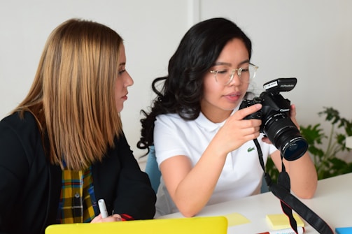 Two people are sitting at a table, engaged in a conversation. One person is holding a professional DSLR camera, and appears to be explaining something about it. The other person is attentively listening, holding a pen and a notepad. The background is minimalistic, and a plant is visible in the distance.