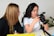 Two people are sitting at a table, engaged in a conversation. One person is holding a professional DSLR camera, and appears to be explaining something about it. The other person is attentively listening, holding a pen and a notepad. The background is minimalistic, and a plant is visible in the distance.