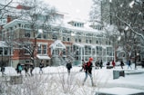 A snowy scene on a college campus with several people walking around. The ground is covered with snow, and trees are bare with snow on their branches. A large building with red brick and white accents dominates the background. Snow is gently falling, and the sky appears overcast.