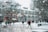 A snowy scene on a college campus with several people walking around. The ground is covered with snow, and trees are bare with snow on their branches. A large building with red brick and white accents dominates the background. Snow is gently falling, and the sky appears overcast.