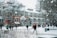 A snowy scene on a college campus with several people walking around. The ground is covered with snow, and trees are bare with snow on their branches. A large building with red brick and white accents dominates the background. Snow is gently falling, and the sky appears overcast.