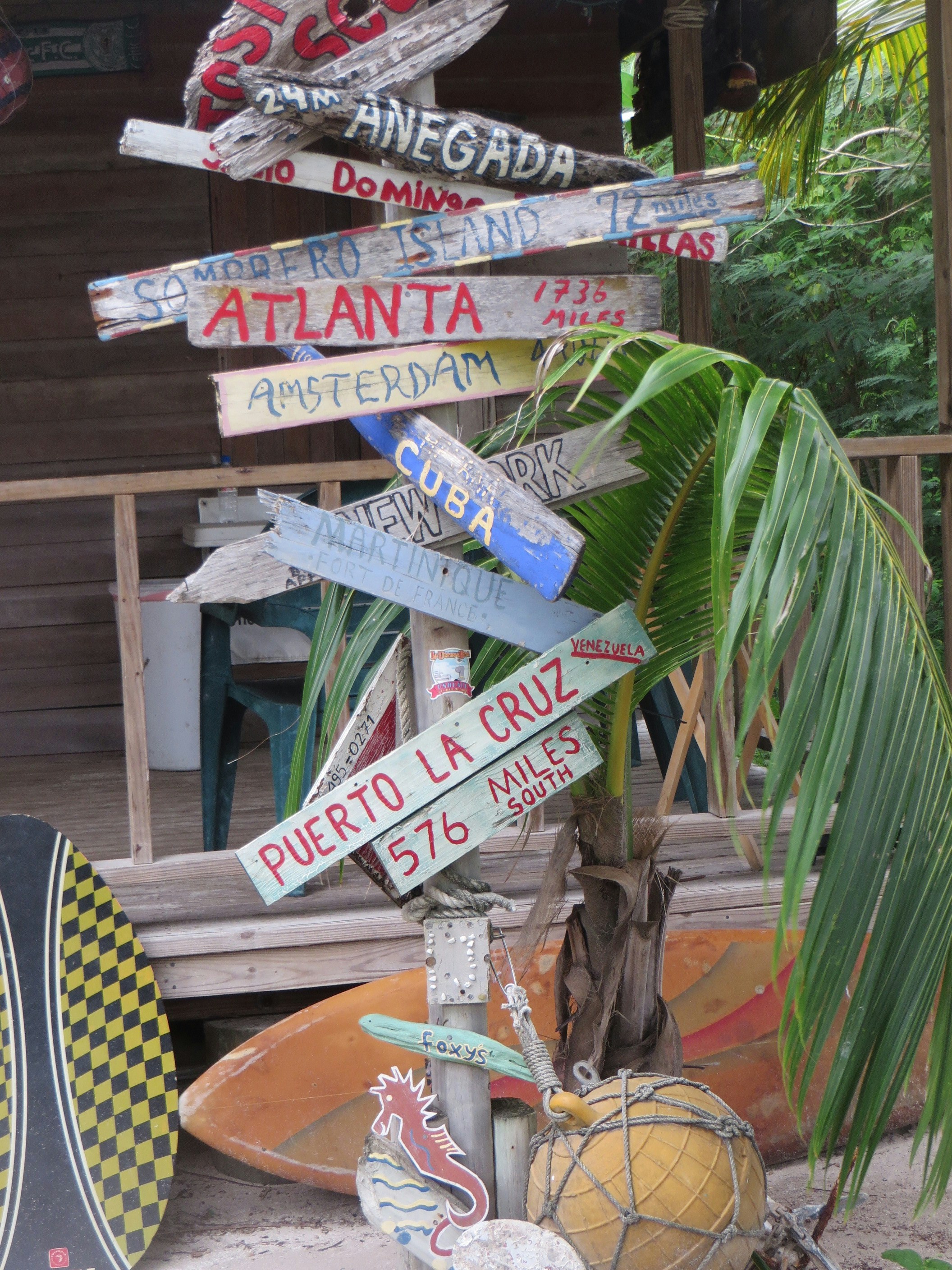 A collection of colorful wooden signs nailed together on a post, each pointing in different directions and displaying names of international destinations such as Atlanta, Puerto La Cruz, Amsterdam, and Cuba, along with the distances in miles. Some palm leaves and nautical objects like a decorative buoy and a seahorse adorn the base. A surfboard and a wooden building with a porch are visible in the background.