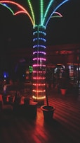 A vibrant, multi-colored LED light installation resembling a palm tree is the focal point of a dimly lit indoor space. The floor is wooden with planters arranged around the base of the light structure, and soft ambient lighting reflects off different surfaces.