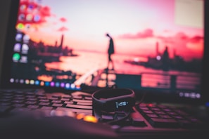A close-up view of a computer keyboard with a fitness tracker resting on top, set against a blurred background displaying a sunset over a cityscape on a monitor.