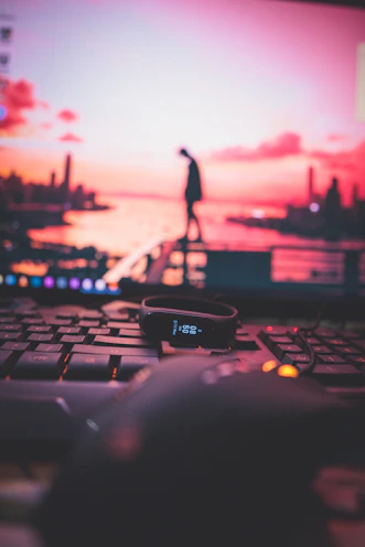 A black fitness tracker displaying the time sits atop a keyboard with a computer mouse nearby. In the background, a blurred computer screen shows a silhouette of a person standing on a railing against a vibrant pink and orange sunset over a cityscape.