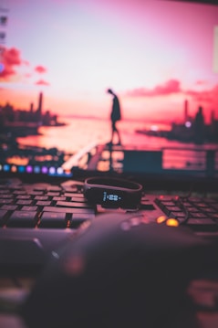 A black fitness tracker displaying the time sits atop a keyboard with a computer mouse nearby. In the background, a blurred computer screen shows a silhouette of a person standing on a railing against a vibrant pink and orange sunset over a cityscape.