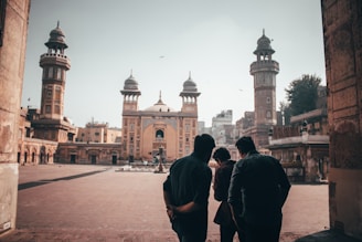A group of people stands in the courtyard of an ornate mosque with tall minarets and intricate architectural details. The building showcases traditional Islamic design, and the scene is lit by soft daylight. The atmosphere appears calm and contemplative.