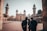 A group of people stands in the courtyard of an ornate mosque with tall minarets and intricate architectural details. The building showcases traditional Islamic design, and the scene is lit by soft daylight. The atmosphere appears calm and contemplative.