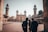 A group of people stands in the courtyard of an ornate mosque with tall minarets and intricate architectural details. The building showcases traditional Islamic design, and the scene is lit by soft daylight. The atmosphere appears calm and contemplative.