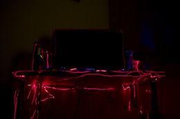 A dark room with glowing red neon or LED lights outlining the edges of a computer monitor and running along a desk. The lights create a striking contrast against the dim surroundings, suggesting a futuristic or gaming atmosphere.