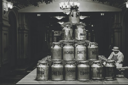A stack of vintage milk cans arranged in a pyramid shape, displayed indoors with decorative straw around them. A person wearing a hat and striped apron is seated at the side, possibly tending to the display. The setting has an old-fashioned, historical ambiance with dim lighting and traditional architectural elements.