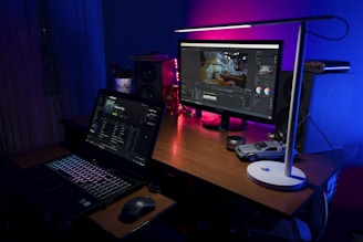 A workspace with a wooden desk featuring a laptop displaying a music streaming application and an external monitor showing video editing software. A sleek white lamp casts light over the area, highlighting items such as a small model car, camera lens caps, a speaker, and colorful light accents creating a cozy atmosphere.