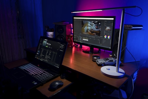 A workspace with a wooden desk featuring a laptop displaying a music streaming application and an external monitor showing video editing software. A sleek white lamp casts light over the area, highlighting items such as a small model car, camera lens caps, a speaker, and colorful light accents creating a cozy atmosphere.