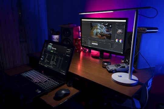 A workspace with a wooden desk featuring a laptop displaying a music streaming application and an external monitor showing video editing software. A sleek white lamp casts light over the area, highlighting items such as a small model car, camera lens caps, a speaker, and colorful light accents creating a cozy atmosphere.