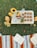 A picnic setup on grass includes a tray with a vase of sunflowers, a bowl of fruits, several oatmeal cookies, and a pitcher of orange juice. Surrounding the tray is a colorful striped blanket with an open notebook and a book. A small succulent and some flowers also decorate the scene.