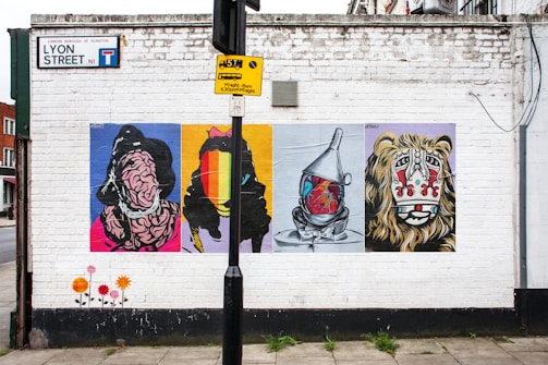 A white brick wall features four colorful street art posters. Each poster depicts a different abstract design, including a distorted figure and a vibrant faceless silhouette. An industrial object resembling a tin man with a heart illustration is portrayed on the third poster, and the fourth features a stylized lion face with tribal patterns. A street sign and a parking restriction notice are present above the artwork. The pavement below includes painted floral designs.