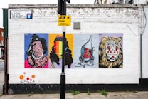 A white brick wall features four colorful street art posters. Each poster depicts a different abstract design, including a distorted figure and a vibrant faceless silhouette. An industrial object resembling a tin man with a heart illustration is portrayed on the third poster, and the fourth features a stylized lion face with tribal patterns. A street sign and a parking restriction notice are present above the artwork. The pavement below includes painted floral designs.