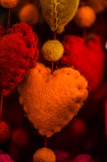 Close-up shot of colorful felt hearts, with a central focus on an orange heart. The background contains various red and green heart-shaped felt decorations, creating a warm and cozy atmosphere.