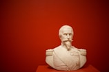 A white marble bust of a man with a mustache and goatee is displayed against a solid red background. The bust appears to be detailed and features a military uniform with epaulettes and several medals embossed on the chest.