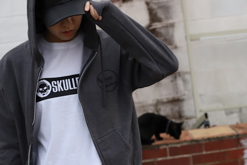 A person is wearing a dark hoodie and a cap, partially covering their face. The white t-shirt underneath has a graphic design with the text 'SKULL' and a skull logo. In the background, a black cat walks along a brick wall, and the wall behind them has a weathered, rough texture.
