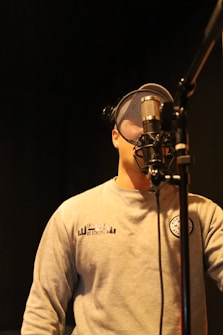 A person wearing a light gray sweatshirt stands behind a large metallic microphone in a recording studio. The background is dimly lit, creating a focused and intimate setting. The person wears a cap with a mesh front, suggesting they might be singing or speaking into the microphone.