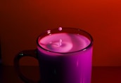 A glass mug filled with milk is placed against a vibrant red backdrop. The milk is slightly disturbed, creating a small ripple effect with a drop captured mid-air just above the surface.