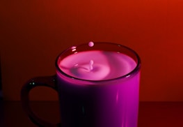 A glass mug filled with milk is placed against a vibrant red backdrop. The milk is slightly disturbed, creating a small ripple effect with a drop captured mid-air just above the surface.