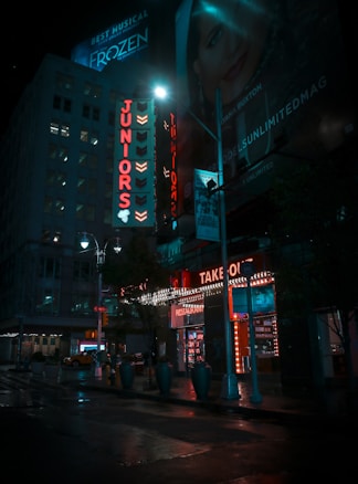 A nighttime street scene featuring brightly lit signage for a restaurant named 'Juniors' in red neon lights. The &lsquo;TAKE OUT&rsquo; section of the restaurant is adorned with lights around the entrance, casting a glow onto the wet pavement. Nearby, a bright billboard advertises for the musical 'Frozen', and another billboard shows a woman's face. The street is relatively quiet with few people visible and a couple of taxis parked along the sidewalk. Tall buildings and street lamps are also present, contributing to the urban atmosphere.