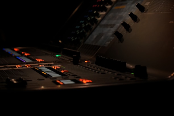 A dimly lit audio mixing console with various knobs, buttons, and sliders illuminated by bright orange, green, and blue lights. The console has a sleek, modern design, and its surface is covered in numerous controls used for sound editing.