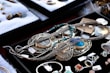 A variety of jewelry pieces are displayed on a black surface. The collection includes intricate necklaces, rings, and pendants with elaborate designs. Most items feature turquoise, silver, or gold elements. Other gemstones and materials are also visible. The surrounding area contains cushioned trays with additional stone pieces and jewelry parts. Each item has a small price tag attached.
