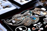 A variety of jewelry pieces are displayed on a black surface. The collection includes intricate necklaces, rings, and pendants with elaborate designs. Most items feature turquoise, silver, or gold elements. Other gemstones and materials are also visible. The surrounding area contains cushioned trays with additional stone pieces and jewelry parts. Each item has a small price tag attached.