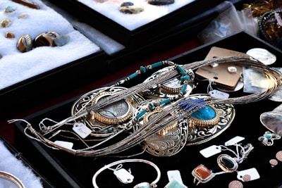A variety of jewelry pieces are displayed on a black surface. The collection includes intricate necklaces, rings, and pendants with elaborate designs. Most items feature turquoise, silver, or gold elements. Other gemstones and materials are also visible. The surrounding area contains cushioned trays with additional stone pieces and jewelry parts. Each item has a small price tag attached.