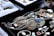 A variety of jewelry pieces are displayed on a black surface. The collection includes intricate necklaces, rings, and pendants with elaborate designs. Most items feature turquoise, silver, or gold elements. Other gemstones and materials are also visible. The surrounding area contains cushioned trays with additional stone pieces and jewelry parts. Each item has a small price tag attached.