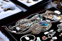 A variety of jewelry pieces are displayed on a black surface. The collection includes intricate necklaces, rings, and pendants with elaborate designs. Most items feature turquoise, silver, or gold elements. Other gemstones and materials are also visible. The surrounding area contains cushioned trays with additional stone pieces and jewelry parts. Each item has a small price tag attached.