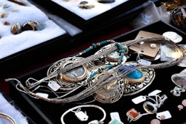 A variety of jewelry pieces are displayed on a black surface. The collection includes intricate necklaces, rings, and pendants with elaborate designs. Most items feature turquoise, silver, or gold elements. Other gemstones and materials are also visible. The surrounding area contains cushioned trays with additional stone pieces and jewelry parts. Each item has a small price tag attached.