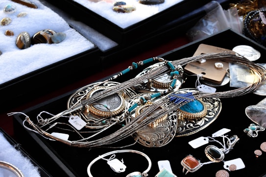 A variety of jewelry pieces are displayed on a black surface. The collection includes intricate necklaces, rings, and pendants with elaborate designs. Most items feature turquoise, silver, or gold elements. Other gemstones and materials are also visible. The surrounding area contains cushioned trays with additional stone pieces and jewelry parts. Each item has a small price tag attached.
