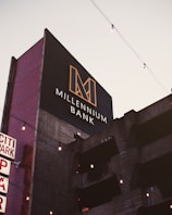 A modern building with a sign reading 'Millennium Bank' features a bold, geometric logo in gold against a dark background. The building facade appears to be concrete with horizontal lines and vertical red brick sides. There are decorative string lights hanging across the scene, and the sky is lightly overcast.
