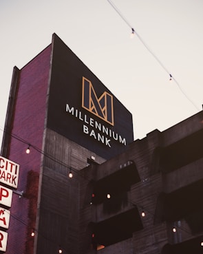 A modern building with a sign reading 'Millennium Bank' features a bold, geometric logo in gold against a dark background. The building facade appears to be concrete with horizontal lines and vertical red brick sides. There are decorative string lights hanging across the scene, and the sky is lightly overcast.