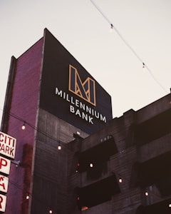 A modern building with a sign reading 'Millennium Bank' features a bold, geometric logo in gold against a dark background. The building facade appears to be concrete with horizontal lines and vertical red brick sides. There are decorative string lights hanging across the scene, and the sky is lightly overcast.