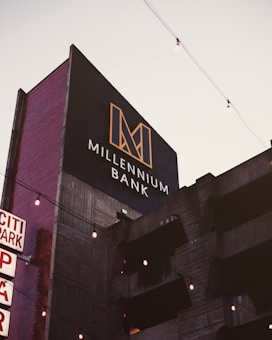 A modern building with a sign reading 'Millennium Bank' features a bold, geometric logo in gold against a dark background. The building facade appears to be concrete with horizontal lines and vertical red brick sides. There are decorative string lights hanging across the scene, and the sky is lightly overcast.
