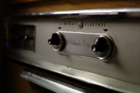 A close-up view of the control panel of a vintage General Electric automatic oven. The panel has a metallic finish with two black knobs and engraved text, surrounded by textured metal.