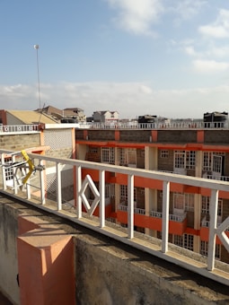 A view from a rooftop showcases residential buildings with multiple stories. The buildings have a combination of beige and orange accents, bordered by a white perimeter railing with geometric designs. In the background, other residential units with sloped roofs are visible under a partly cloudy sky.