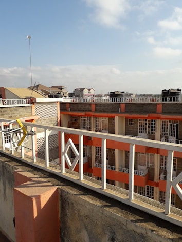 A view from a rooftop showcases residential buildings with multiple stories. The buildings have a combination of beige and orange accents, bordered by a white perimeter railing with geometric designs. In the background, other residential units with sloped roofs are visible under a partly cloudy sky.