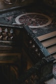 An intricately designed marble staircase with ornate balustrades is positioned against a decorative, circular floor pattern made of dark and light marble. The area appears dimly lit, creating a dramatic contrast with shadows and highlights.