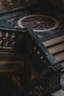 An intricately designed marble staircase with ornate balustrades is positioned against a decorative, circular floor pattern made of dark and light marble. The area appears dimly lit, creating a dramatic contrast with shadows and highlights.