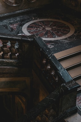 An intricately designed marble staircase with ornate balustrades is positioned against a decorative, circular floor pattern made of dark and light marble. The area appears dimly lit, creating a dramatic contrast with shadows and highlights.