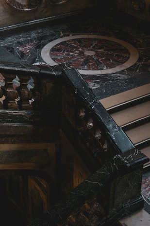 An intricately designed marble staircase with ornate balustrades is positioned against a decorative, circular floor pattern made of dark and light marble. The area appears dimly lit, creating a dramatic contrast with shadows and highlights.