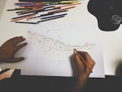 A person is drawing a detailed geological or topographical sketch on white paper. Various colored pencils are scattered above the paper on a white desk. The sketch appears to depict a cross-section of land with annotations.